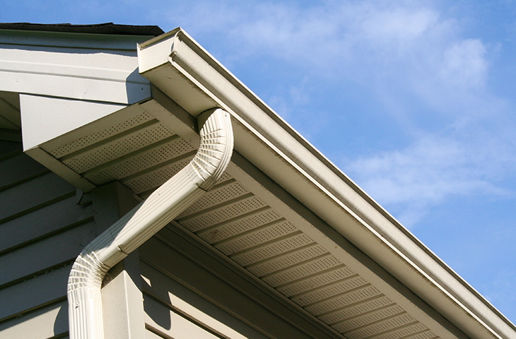 A close-up view of a house’s roofline shows a white gutter system and downspout against blue sky, with vented soffit panels and beige siding visible.