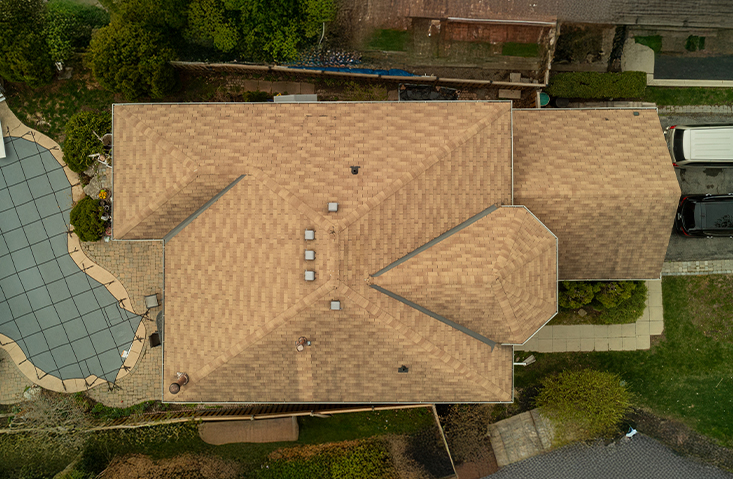Aerial view of a tan shingle roof on a suburban house, surrounded by trees, a driveway with vehicles, a covered swimming pool, and a sidewalk leading to the entrance.
