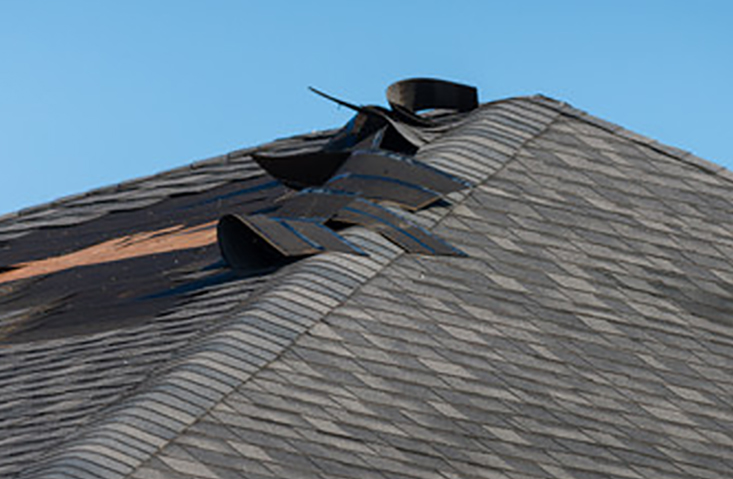 A shingled roof with several shingles peeled back and damaged, exposing the underlying structure, under a clear blue sky.