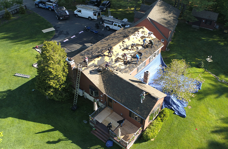 Aerial view of several workers replacing the roof of a house, with tools and materials scattered on the roof and ground. Ladders, trucks, and tarps are visible around the house, which is surrounded by green lawn and trees.