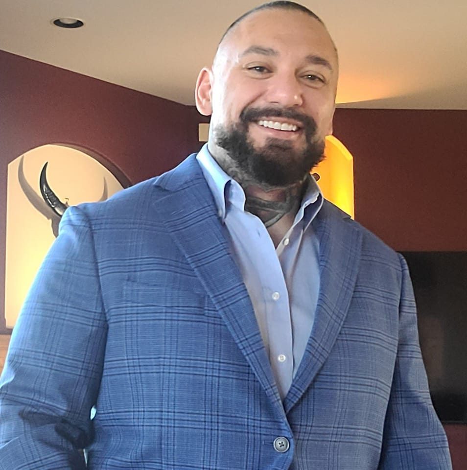 A smiling man with a beard and tattoos on his neck is wearing a blue plaid suit jacket and light blue shirt, standing indoors in a warmly lit room.